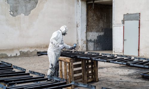 The painter in protective clothing, paints metal products in the open air, using a spray gun. The picture was taken in Russia, in the city of Orenburg, on the territory of an industrial enterprise